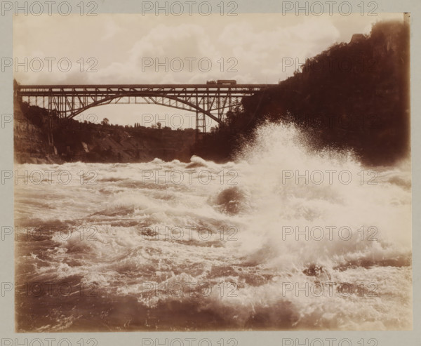 Bridge between America and Canada, United States, Canada, photograph, Bridge between America and Canada seen from the water., annotation: '10. Bridge between Canada & America', Niagara Falls (North America), c. 1895 - c. 1915, photographer, North America, paper, pasteboard, albumen print, height 193 mm x width 243 mm, height 243 mm x width 329 mm, bridge, A wide sepia-toned photograph showing turbulent water in the foreground with frothy waves and spray rising, a large spray plume toward the right side, and a steel arch bridge spanning the scene in the midground with visible trusses and supports; a small rail car sits on the bridge, cliffs and tree-covered slopes flank both sides of the river, and cloudy sky fills the upper portion of the image.