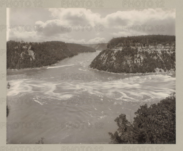Whirlpool at Niagara Falls, United States, Canada, photograph, annotation: '11. Whirlpool.', Niagara Falls (North America), c. 1895 - c. 1915, photographer, North America, paper, pasteboard, gelatin silver print, developing gelatin silver print, height 192 mm x width 240 mm, height 243 mm x width 329 mm, A wide river flows between steep forested cliffs with swirling lighter patches on the water surface and rocky shorelines, dense trees covering the slopes and tops of the cliffs, a partly cloudy sky with large billowy clouds above, and foliage visible in the lower foreground along the river edge.