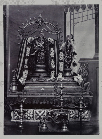 Sculptures for worship of Shiva-Nataraja and Parvati inside the Meenakshi temple at Madurai, Tamil Nadu, India, photograph, annotation: '77b. Temple at Madura.', Madurai, c. 1895 - c. 1915, photographer, South India, pasteboard, paper, daylight gelatin silver print, height 205 mm x width 151 mm, height 244 mm x width 164 mm, Person, Shiva, Parvati, A central ornate shrine with two decorated statues is shown on a raised platform; the larger statue on the left is seated within a circular halo adorned with small floral motifs and draped with patterned cloth and garlands of flowers, while the smaller statue on the right stands on a pedestal wearing a tall headdress and multiple garlands. The platform features carved floral panels and steps, with several metal oil lamps and a curved metal vessel arranged in front. The background includes a painted architectural scene with a column and an archway, and the entire composition is monochrome with varied textures of metal, fabric, and fresh flora...