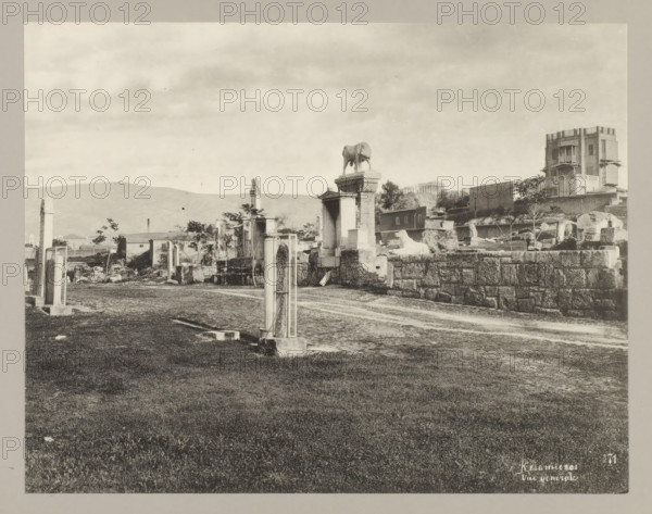 Keramiecos Vue generale 71, View of Kerameikos, photograph, annotation: '31. Cemetery at Athens.', Athens, c. 1895 - c. 1915, photographer, Athene, Periféreia Protevoúsis, pasteboard, paper, collotype, photoprint, height 215 mm x width 276 mm, height 239 mm x width 328 mm, A wide, monochrome scene showing an open grassy foreground leading to a dirt path and a ruined stone area with scattered columns and blocks. Along the path are upright stone posts and rectangular framed structures, and further back are larger stone pedestals and sculpted forms including a statue of an animal standing on a plinth and a reclining animal figure on a raised stone platform. Behind these ruins are low buildings, partial walls, and a taller multi-story building with balconies and decorative openings. In the distant background low hills are visible beneath a cloudy sky.