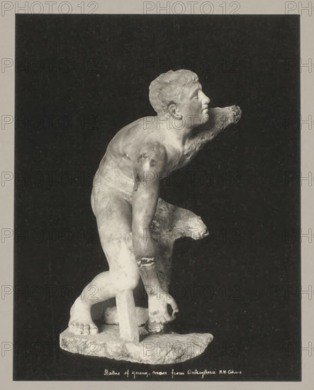 Statue of young man from Anticythera NM Athens, Statue of young man from Antikythera, photograph, number: '34.', Athens, c. 1895 - c. 1915, photographer, Athene, Periféreia Protevoúsis, pasteboard, paper, collotype, photoprint, height 274 mm x width 215 mm, height 328 mm x width 239 mm, A grayscale photograph of a partially damaged classical marble statue of a young male figure set against a dark rectangular background within a lighter border, showing the figure leaning forward with one arm extended and the other bent, a supportive tree-stump-like element behind one leg, detailed musculature and curly hair on the head turned upward, visible feet standing on a rough rectangular base, and areas of surface wear and missing fragments on the arms and torso.