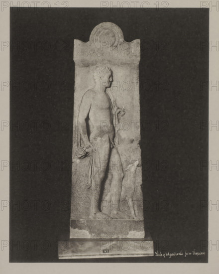 Stèle of Thepiae, Stele of Agathocles from Thepiae, photograph, number: '38.', Athens (possibly), c. 1895 - c. 1915, photographer, Athene, Periféreia Protevoúsis, pasteboard, paper, collotype, photoprint, height 273 mm x width 273 mm, height 328 mm x width 239 mm, A tall rectangular stone relief stands against a dark rectangular backdrop, showing a carved male figure in profile facing right with short curly hair and a draped garment hanging from one shoulder and arm, the figure's bare torso and legs visible and one hand holding a small object while the other hand rests near the hip, a dog-like animal stands beside the figure with its head raised, the relief has a rounded decorative top and rests on a plinth with a small rectangular plaque, the surface of the stone shows weathered texture and worn edges.