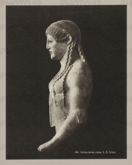 686 Archaic female statue. AM Athens, Archaic statue of a woman, photograph, number: '39.', Athens, c. 1895 - c. 1915, photographer, Athene, Periféreia Protevoúsis, pasteboard, paper, collotype, photoprint, height 274 mm x width 215 mm, height 328 mm x width 239 mm, A grayscale photograph shows a side view of a classical female statue against a dark rectangular background framed by a lighter border, depicting the statue from head to forearm; the figure has stylized wavy hair pulled back and falling in long undulating locks, a smooth profile with a straight nose and closed lips, a draped garment with vertical folds and decorative banding over the chest, a rounded shoulder and upper arm with some surface irregularities, and an extended forearm ending in a partially visible hand.