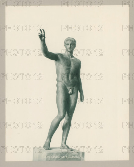 13396. Ephebus from Anticythera., Statue of Ephebus from Antikythera, photograph, number: '40.', Athens, c. 1895 - c. 1915, photographer, Athene, Periféreia Protevoúsis, pasteboard, paper, collotype, photoprint, height 273 mm x width 217 mm, height 328 mm x width 239 mm, A full-length photograph of a classical male nude statue standing on a rectangular plinth, shown against a plain light background and framed by a narrow darker border, with the figure posed with one arm raised and the other relaxed by the side, curly hair, defined musculature, and both feet placed on the plinth.