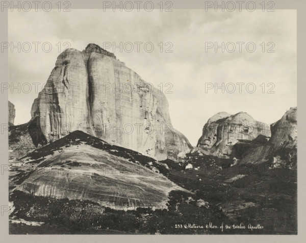 238 Meteora Mon. of the twelve Apostles, View of Meteora, photograph, number: '46.', unknown, c. 1895 - c. 1915, photographer, pasteboard, paper, collotype, photoprint, height 215 mm x width 275 mm, height 239 mm x width 328 mm, Thessaly, A wide photographic view shows large, smooth rock formations rising from a rugged landscape, with one prominent vertical face occupying the left-center and other rounded cliffs to the right; the rock surfaces display striations, grooves, and lighter and darker patches, and scattered vegetation clings to slopes and crevices, while the foreground contains uneven rocky ground and terraced or patterned patches of lighter texture; the sky above is mostly pale and cloudy.