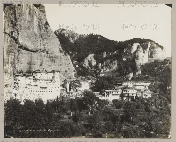 78 Vue générale du monastère de Megaspiléon, View of the monastery of Megaspileon, photograph, number: '48.', unknown, c. 1895 - c. 1915, photographer, pasteboard, paper, gelatin silver print, developing gelatin silver print, height 224 mm x width 278 mm, height 239 mm x width 328 mm, Thessaly, A black and white photograph showing a steep rocky cliff face on the left and a rugged hillside with rocky outcrops and dense tree cover occupying the center and right, with multi-story buildings of varying sizes and shapes built along the lower slopes and at the base of the cliffs, some structures appearing terraced into the rock; the foreground contains darker trees and vegetation, and the upper portion of the image shows an expanse of pale sky.