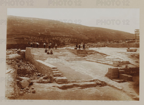 Palace of Knossos, photograph, annotation: '50. Crete, palace of Cnossos.', unknown, c. 1895 - c. 1915, photographer, pasteboard, paper, daylight gelatin silver print, height 155 mm x width 226 mm, height 239 mm x width 328 mm, Crete, Knossós, A wide sepia-toned scene of an archaeological site with broad stone terraces and stepped platforms leading up to a higher level where a group of people stand and sit; scattered stone blocks and rubble lie in the foreground and along a sunken trench with wooden beams, while low stone walls and roughly hewn blocks form retaining structures across the site; a hillside with sparse vegetation rises in the background under a pale sky, and a wooden scaffold-like structure stands near the right edge of the image.