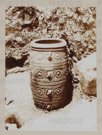 Large vase (?) at the palace of Knossos, photograph, annotation: '54. Crete. Palace at Cnossos.', unknown, c. 1895 - c. 1915, photographer, pasteboard, paper, daylight gelatin silver print, height 219 mm x width 162 mm, height 328 mm x width 239 mm, Crete, Knossós, A tall cylindrical ceramic vessel sits upright on a stone surface against a rough rock wall, the vessel featuring multiple horizontal bands of decorative raised patterns including circular rosettes, beaded rows, and twisted rope-like motifs, with protruding rounded knobs at intervals and a wide rounded rim at the top; the surrounding area shows uneven rocky textures and light and shadow contrasts on the ground and wall.