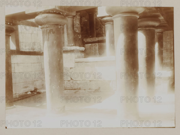 Wooden columns in the Palace of Knossos, photograph, annotation: '55. Crete. Palace at Knossos (Wooden columns: restored).', unknown, c. 1895 - c. 1915, photographer, pasteboard, paper, daylight gelatin silver print, height 163 mm x width 228 mm, height 239 mm x width 328 mm, Crete, Knossos, A sepia-toned photograph showing a row of large rounded columns with bulbous capitals supporting horizontal beams, a stone wall with visible block joints behind the columns, a raised stone ledge and recessed areas between the columns, worn and patched surfaces on the columns and walls, shadowed floor area beneath the columns, and bright overexposed light in the foreground that fades upward.