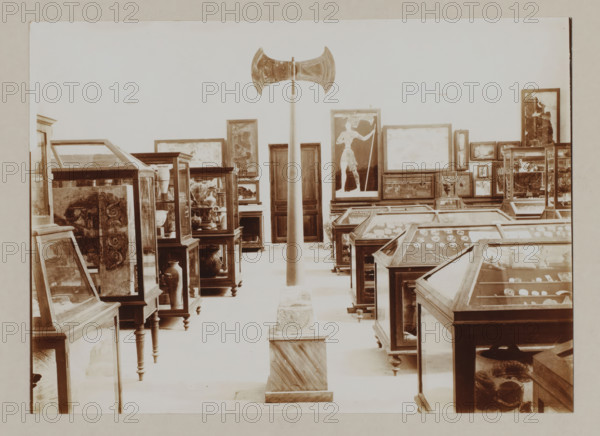 Double axe in museum on Crete, photograph, Double axe, symbol of divinity, amidst cabinets with objects., annotation: '57. Crete. Museum at Candia Double axe symbol of divinity', unknown, c. 1895 - c. 1915, photographer, pasteboard, paper, daylight gelatin silver print, height 166 mm x width 228 mm, height 239 mm x width 328 mm, A large indoor space filled with glass display cases and framed artworks is shown, with a tall vertical pole topped by a double-bladed axe positioned centrally and mounted on a square plinth at floor level, wooden-framed glass cases arranged in rows on either side containing various objects and pottery, framed paintings and panels leaning against the walls and stacked on furniture, one prominent framed image of a standing figure near the center right, wooden cabinets and pedestals supporting many of the displays, and a pale wall forming the backdrop.