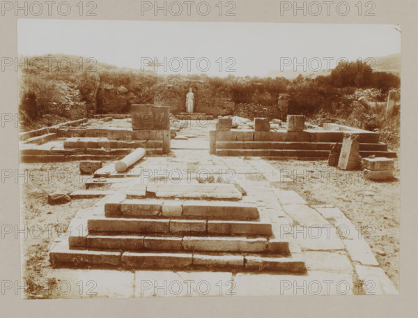Temple of Apollo at Phaestus, Crete, photograph, annotation: '63 Crete. Phestos, Apollo temple at Phestos.', unknown, c. 1895 - c. 1915, photographer, pasteboard, paper, daylight gelatin silver print, height 160 mm x width 224 mm, height 239 mm x width 328 mm, A sepia-toned photograph shows an archaeological site with a stepped stone platform leading toward a rectangular paved area scattered with stone blocks and fallen columns, flanked by low stone walls and raised stone foundations; at the far end a freestanding statue is positioned against a short brick wall surrounded by vegetation and low hills, with bushes and trees visible along the edges and broken stone fragments and blocks resting on the ground.