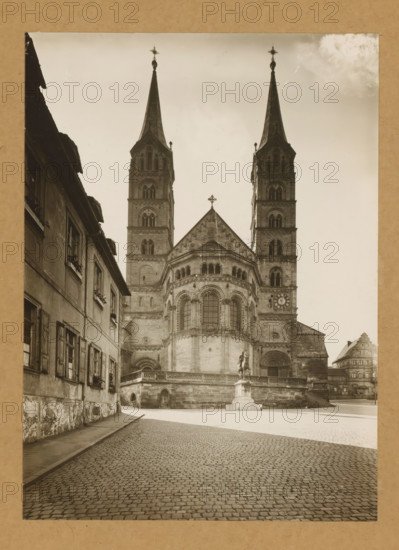Ostansicht, View of Bamberg Cathedral, album page, photograph, This photo is part of an album., number: '1', Bamberg, 1900 - c. 1920, photographer, Bavaria, paper, baryta paper, gelatin silver print, developed gelatin silver print, height 227 mm x width 165 mm, church (exterior), Bamberg Cathedral, A stone cathedral facade with twin tall spires topped by crosses rises from a raised platform accessed by steps, its central section featuring rounded arches and a large arched window flanked by decorative columns, while a statue on a pedestal stands in front of the platform; a cobblestone street leads toward the building, bordered on the left by an old plastered row of buildings with shuttered windows and flower boxes, and on the right by additional stone structures partially visible in the distance, all shown in a sepia-toned photograph.