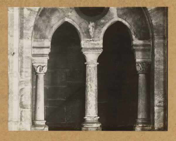 Eastern choir, inner window, View of a window of Bamberg Cathedral, album page, photograph, This photo is part of an album., number: '10', Bamberg, 1900 - c. 1920, photographer, Bavaria, paper, baryta paper, gelatin silver print, developed gelatin silver print, height 168 mm x width 217 mm, church (exterior), Bamberg Cathedral, A close view of a stone architectural opening featuring three arched openings supported by rounded columns with molded capitals and bases, the stone showing weathered textures and uneven coloration, decorative carved leaf motifs on the outer capitals, a dark recessed background visible through the arches, and surrounding vertical stone jambs with worn surfaces.