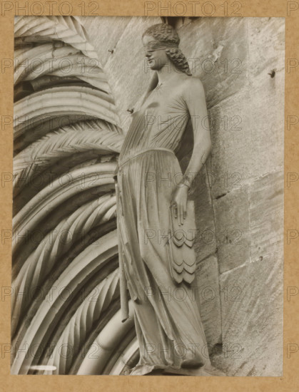 Detail of the Fürstenportal in Bamberg Cathedral, Fürstentor, Synagogue v. Halbrechts. 106a, dasselbe, album page, photograph, This photo is part of an album., number: '106.', Bamberg, 1900 - c. 1920, photographer, Bavaria, paper, baryta paper, gelatin silver print, developing gelatin silver print, height 211 mm x width 160 mm, church (exterior), Bamberg Cathedral, A grayscale photograph of a sculpted female figure set against a textured stone background, the figure wearing a long draped gown with visible folds and a belted waist, a blindfold covering the eyes, wavy hair pulled back, one arm hanging by the side with a layered decorative element resembling feathers, and the other arm partly visible; to the left of the figure a series of rounded, ribbed, and braided architectural carvings form a repeating pattern that fills the background.