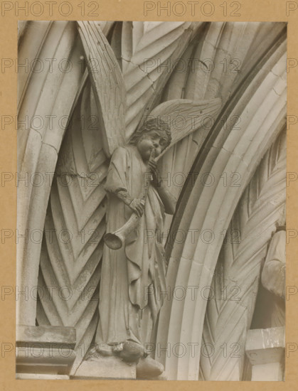 Detail of the Princely Portal in Bamberg Cathedral, Princely Gate, Synagogue, Posaunenengel vl, album page, photograph, This photo is part of an album., number: '107.', Bamberg, 1900 - c. 1920, photographer, Bavaria, paper, baryta paper, gelatin silver print, gelatin silver developing print, height 217 mm x width 163 mm, church (exterior), Bamberg Cathedral, A stone carving of an angel with textured wings stands within a series of arched, ribbed stone surfaces; the angel wears flowing robes with visible folds and holds a long trumpet to its lips while one hand supports the instrument, the face showing carved features and wavy hair; surrounding the figure are layered, chevron-patterned and fluted stone arches with weathered surfaces and small chips and cracks, all shown in a monochrome tone against a plain border.