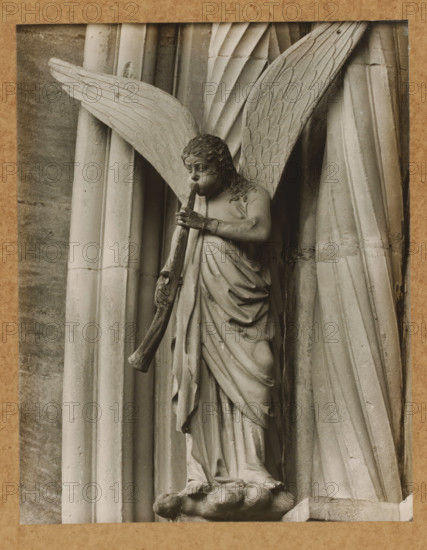 Detail of the Princely Portal in Bamberg Cathedral, Princely Gate, Trombone Angel from right, album page, photograph, This photo is part of an album., number: '108', Bamberg, 1900 - c. 1920, photographer, Bavaria, paper, baryta paper, gelatin silver print, height 207 mm x width 162 mm, church (exterior), Bamberg Cathedral, A carved stone figure of an angel is shown against a background of vertical fluted columns, with large feathered wings extending outward behind the figure; the angel has wavy hair, a contemplative facial expression, and holds a long curved horn to its mouth with both hands, wearing draped robes that fall in folds to a textured base beneath its feet.