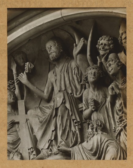Fürstentor, Tymphanon, Christ the Lady, Detail of the tympanum of the Fürstenportal in Bamberg Cathedral, album page, photograph, This photo is part of an album., number: '115', Bamberg, 1900 - c. 1920, photographer, Bavaria, paper, baryta paper, gelatin silver print, gelatin silver developing print, height 208 mm x width 163 mm, church (exterior), Bamberg Cathedral, A close-up photograph of a carved stone relief framed by a textured border, showing multiple sculpted figures in draped robes and detailed hair. One central seated figure has a beard and raised arm with an open hand, while surrounding figures display varied facial expressions and gestures, including a smiling figure with stylized curls and a youthful figure looking upward. Folded and flowing drapery covers limbs and torsos, and carved architectural arches appear above the group. A partial cross is visible and several hands are clasped or holding small objects, with a smaller figure kneeling at the lower edge. The stone sur...