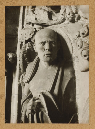 Georgenchorschranken 3 Pvli.v.vorn, Detail of the rood screen in the East Choir of Bamberg Cathedral, album page, photograph, This photo is part of an album., number: '119.', Bamberg, 1900 - c. 1920, photographer, Bavaria, paper, baryta paper, gelatin silver print, developed gelatin silver print, height 224 mm x width 160 mm, interior of church, Bamberg Cathedral, A close view of a carved stone figure shown from the waist up, featuring a bald head with neutral facial expression and a circular halo behind the head, draped cloth over one shoulder, and one hand gripping a bundle of rounded rope-like forms; decorative carved foliage and scroll motifs surround the figure on both sides, and a partial profile of another carved face appears at the right edge.
