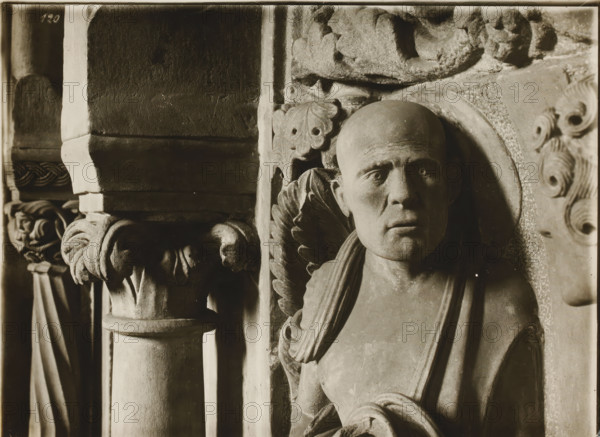 Georgenchorschranken derselbe m. Kapitellen, Detail of the rood loft in the Ostchor of Bamberg Cathedral, album page, photograph, This photo is part of an album., inscription: 'Kunsthist. Seminar, Marburg (Lahn), Photogr. Abteilung, Vervielfältigung, untersagt.', number: 'Bamberg Jonas ?, Bamberg, 120', 1900 - c. 1920, photographer, Bavaria, paper, baryta paper, gelatin silver print, developing gelatin silver print, height 180 mm x width 239 mm, interior of church, Bamberg Cathedral, A close-up sepia photograph shows a stone relief of a bald human head and upper torso with a solemn facial expression and deep-set eyes, flanked by carved decorative foliage and a curved halo behind the head; to the left a textured column with a carved capital featuring rounded, folded forms and a small sculpted head is visible, and further left vertical fluted elements and draped carved fabric appear; the right edge shows part of another carved face with stylized hair.