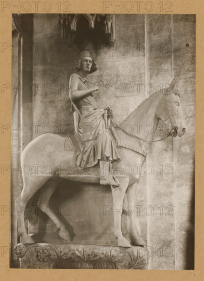 Bamberger Reiter in the cathedral of Bamberg, Der Reiter, Knitting page, album page, photograph, This photo is part of an album., number: '131.', Bamberg, 1900 - c. 1920, photographer, Bavaria, paper, baryta paper, gelatin silver print, developed gelatin silver print, height 222 mm x width 160 mm, riding a horse, ass, or mule, rider, horseman, interior of church, Cathedral of Bamberg, A stone equestrian statue occupying most of the frame shows a robed rider seated on a bridled horse, both carved with detailed drapery and tack; the rider wears a crown-like headpiece and has curly hair, a cloak draped over one shoulder, and one hand holding a strap across the chest while the other rests near the waist, the folds of the robe falling over the horse's flank and legs; the horse stands with a raised head, visible bridle and bit, and textured mane, its legs positioned on a carved rectangular plinth decorated with acanthus leaves and floral motifs; the background consists of a stone wall with v...