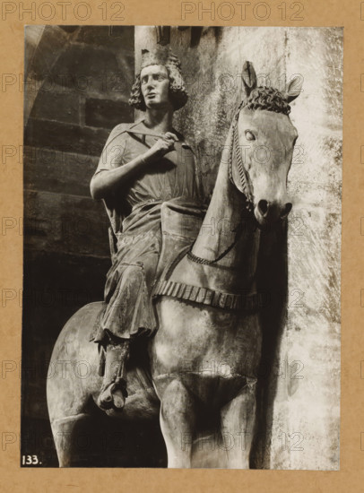 The rider, from the front, Bamberg horseman in the cathedral of Bamberg, album page, photograph, This photo is part of an album., number: '133.', Bamberg, 1900 - c. 1920, photographer, Bavaria, paper, baryta paper, gelatin silver print, developing gelatin silver print, height 224 mm x width 161 mm, riding a horse, ass, or mule, rider, horseman, interior of church, Cathedral of Bamberg, A monochrome photograph of a stone equestrian statue showing a rider with curly hair and a crown or headpiece seated on a horse, the rider wearing a draped garment and holding a strap or rein near the chest, the horse facing forward with detailed bridle and a short mane, the sculpture positioned against a textured wall and framed by a darker background on one side, the image bordered by a plain mat.