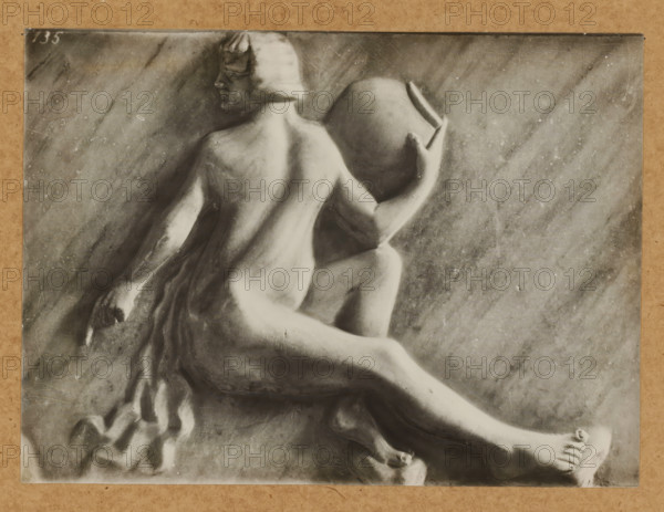 Grabmal, Prudentia, Detail of the tomb of Pope Clement II in the cathedral of Bamberg, album page, photograph, This photo is part of an album., number: '135', Bamberg, 1900 - c. 1920, photographer, Bavaria, paper, baryta paper, gelatin silver print, developing gelatin silver print, height 166 mm x width 226 mm, Person, Clemens II, interior of church, Bamberg Cathedral, A grayscale photograph of a sculpted relief showing a seated nude figure viewed from behind and in profile, with a bobbed hairstyle and turned head, one arm bent and holding a large round vessel against the shoulder while the other arm rests downward, draped fabric cascading beside the figure, a textured stone background with diagonal streaking and surface blemishes, and the image bordered by a light brown mat.
