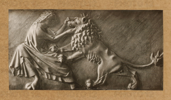 Detail of the tomb of Pope Clement II in the cathedral of Bamberg, Grabmal, Stärke, album page, photograph, This photo is part of an album., number: '140', Bamberg, 1900 - c. 1920, photographer, Bavaria, paper, baryta paper, gelatin silver print, developing gelatin silver print, height 115 mm x width 220 mm, Person, Clemens II, interior of church, Bamberg Cathedral, A rectangular relief panel set against a textured border depicts a seated person with long wavy hair and draped clothing facing a large lion; the person’s arms reach toward the lion’s head and the lion rears with a flowing mane and open mouth, its front paws raised and hind legs braced, with detailed musculature and fur textures carved across both figures and a smooth, slightly scratched background.