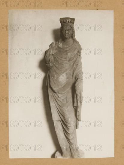 Sculpture in the cloister of Bamberg Cathedral, Cloister of St. Catherine vv, album page, photograph, This photo is part of an album., number: '143.', Bamberg, 1900 - c. 1920, photographer, Bavaria, paper, baryta paper, gelatin silver print, developed gelatin silver print, height 227 mm x width 161 mm, interior of church, Bamberg Cathedral, A grayscale photograph of a standing stone statue depicting a crowned figure draped in flowing robes, shown from head to feet, with a serene facial expression and long wavy hair; the figure holds a small animal against the chest with one hand while the other hand rests by the side, and the statue casts a soft shadow on a plain light background framed by a textured neutral border.