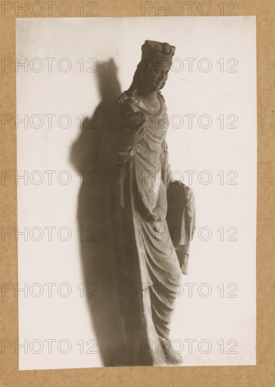 Cloister of St. Catherine of the People, Sculpture in the cloister of Bamberg Cathedral, album page, photograph, This photo is part of an album., number: '144', Bamberg, 1900 - c. 1920, photographer, Bavaria, paper, baryta paper, gelatin silver print, developed gelatin silver print, height 230 mm x width 157 mm, interior of church, Bamberg Cathedral, A grayscale photograph of a draped statue showing a standing figure wearing a crown and layered robes, the figure's head slightly tilted and one arm bent at the elbow while the other draped hand holds fabric, with a strong shadow cast to the left on a pale background and a textured border around the image.