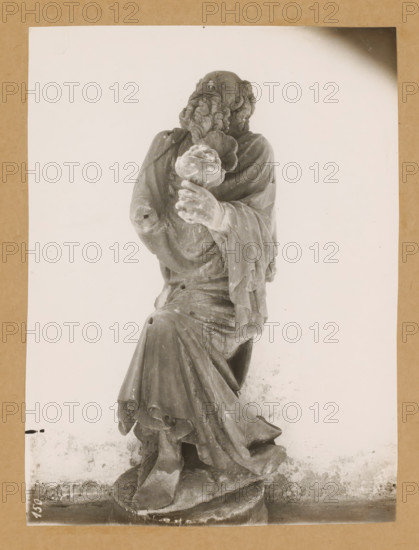 Sculpture in the cloister of Bamberg Cathedral, Cloister, King, from the front, album page, photograph, This photo is part of an album., number: '150', Bamberg, 1900 - c. 1920, photographer, Bavaria, paper, baryta paper, gelatin silver print, developed gelatin silver print, height 228 mm x width 170 mm, interior of church, Bamberg Cathedral, A grayscale photograph of a seated stone statue mounted on a neutral background, showing a robed bearded figure with detailed curly hair and a contemplative upward gaze, one hand raised holding a rounded object with textured surface and the other arm draped by flowing fabric, the robe exhibiting deep folds and drapery that cascade to a carved base with visible wear and surface spots, framed by a rectangular lighter area against a darker textured border.