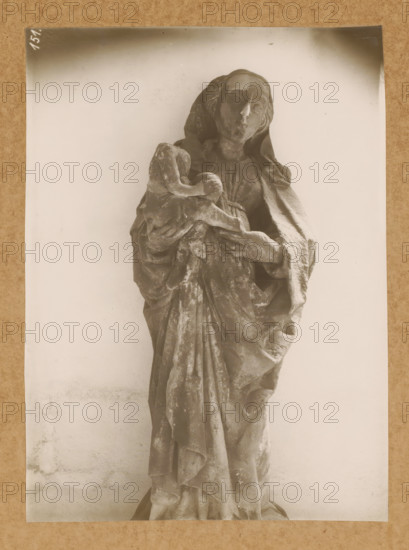 Cloister, Madonna, Sculpture in the cloister of Bamberg Cathedral, album page, photograph, This photo is part of an album., number: '151.', Bamberg, 1900 - c. 1920, photographer, Bavaria, paper, baryta paper, gelatin silver print, height 228 mm x width 168 mm, interior of church, Bamberg Cathedral, A monochrome photograph of a carved figure draped in flowing robes, shown from head to lower torso, standing against a plain light background. The figure has a serene facial expression with closed or lowered eyes and hair partially covered by a hood or veil. One arm cradles a smaller wrapped form while the other supports it, with detailed folds and texture in the garments visible. The surface of the carving shows wear and roughness, and the image is bordered by a narrow darker edge against a textured mat.