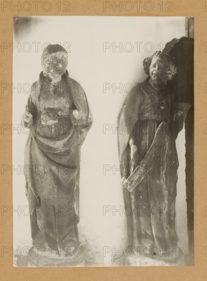 Cloister, Saint (Reverence), Sculpture in the cloister of Bamberg Cathedral, album page, photograph, This photo is part of an album., number: '152', Bamberg, 1900 - c. 1920, photographer, Bavaria, paper, baryta paper, gelatin silver print, height 225 mm x width 164 mm, interior of church, Bamberg Cathedral, Two weathered stone statues stand against a pale background, each mounted on a round base and framed by a neutral border. The statue on the left depicts a robed figure with a hooded or draped garment, hands positioned near the midsection, and a rounded, worn face with indistinct features; the robe shows layered folds and surface erosion. The statue on the right shows a robed figure with curly hair, a tilted head, and a long flowing garment; one arm is bent holding a long curved object while the other arm hangs by the side, and the surface displays chips, scratches, and areas of wear. Both statues exhibit aged textures, weathering marks, and variations in tonal contrast.