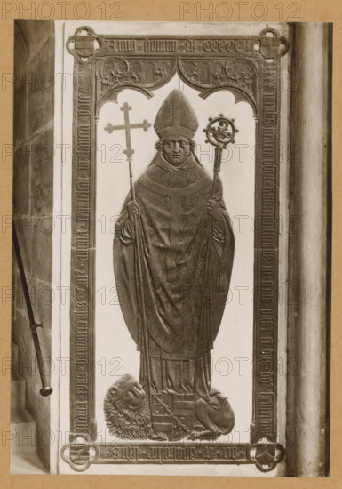 Grave of Heinrich III Groß von Trockau in the cathedral of Bamberg, Heinrich III. Gross v. Trockau (1501) vPVischer, album page, photograph, This photo is part of an album., number: '162', Bamberg, 1900 - c. 1920, photographer, Bavaria, paper, baryta paper, gelatin silver print, developing gelatin silver print, height 225 mm x width 158 mm, Person, Heinrich III. Groß von Trockau, interior of church, Bamberg Cathedral, A framed stone relief shows a robed bishop standing frontally, wearing a tall pointed mitre and ornate vestments with patterned detailing, holding a long staff with a circular ornamental top in one hand and a long cross in the other; his facial features are solemn with curled hair visible at the sides and his hands grasp the staffs; the figure is set within a carved architectural border with decorative arches and floral motifs, and a carved lion lies at the bishop’s feet with a shield resting against it; the relief is monochrome and mounted against a lighter background wi...