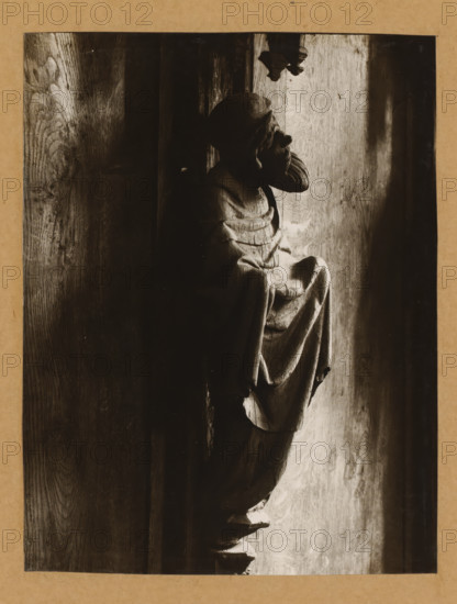 Detail of the choir stalls in Bamberg Cathedral, Chorgestühl i. Peterschor, (Heiliger), album page, photograph, This photo is part of an album., number: '169', Bamberg, 1900 - c. 1920, photographer, Bavaria, paper, baryta paper, gelatin silver print, developed gelatin silver print, height 215 mm x width 164 mm, interior of church, Bamberg Cathedral, A dark-toned photograph of a carved wooden figure shown in profile, positioned against a textured wooden surface; the figure wears a draped garment with visible folds and a head covering, and the carved facial features include a pronounced nose and beard, while light falls from the right side creating highlights and deep shadows across the figure and the wood grain.