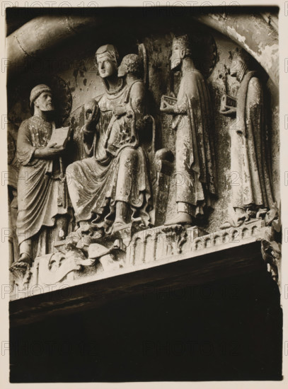 Marienpforte, r. Halfway from the Seite, Detail of the tympanum of the Gnadenpforte in the cathedral of Bamberg, album page, photograph, This photo is part of an album., inscription: 'Kunsthist. Seminar, Marburg (Lahn), Photogr. Abteilung, Vervielfältigung, untersagt.', Bamberg, 1900 - c. 1920, photographer, Bavaria, paper, baryta paper, gelatin silver print, developing gelatin silver print, height 216 mm x width 161 mm, church (exterior), Bamberg Cathedral, A stone relief showing five standing and seated human figures in draped robes, each with distinct headgear and facial features, arranged across a ledge beneath an arch; one central seated figure holds a spherical object while another seated figure beside them leans close, two standing figures to the right present small box-like objects, and a standing figure to the left holds an open rectangular object; the carved surfaces show weathering and textured patina, and decorative carved elements and architectural molding appear along the...