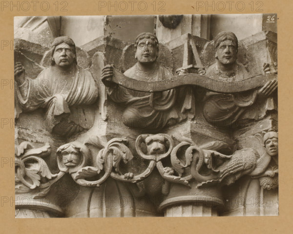 Marienpforte, left. Chapter zone middle from north-east., Detail of a capital of the Gnadenpforte in Bamberg Cathedral, album page, photograph, This photo is part of an album., number: '26.', Bamberg, 1900 - c. 1920, photographer, Bavaria, paper, baryta paper, gelatin silver print, developed gelatin silver print, height 169 mm x width 220 mm, church (exterior), Bamberg Cathedral, A stone relief showing three robed figures in a row holding a curved band, each with distinct facial features and hairstyles, a central figure with a beard and two flanking figures with shorter hair; below them an ornate carved frieze with foliate scrolls, a small human head with stylized hair and a central carved animal face surrounded by curved decorative elements, and a partially visible kneeling figure on the right holding part of the scrollwork; the stone surface shows weathering and small chips, and the composition is set against a plain rectangular backing.