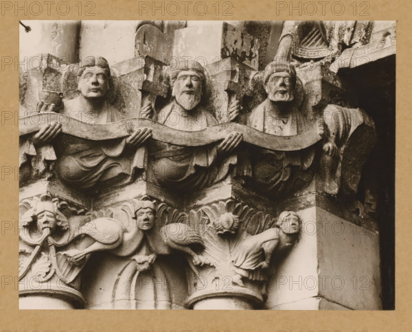 Marienpforte, left. Capital zone Right front, Detail of a capital of the Gnadenpforte in Bamberg Cathedral, album page, photograph, This photo is part of an album., number: '27.', Bamberg, 1900 - c. 1920, photographer, Bavaria, paper, baryta paper, gelatin silver print, developed gelatin silver print, height 168 mm x width 219 mm, church (exterior), Bamberg Cathedral, Stone sculpted figures arranged horizontally, each with distinct facial features and hair, holding a wavy band across their chests; they wear draped garments and one holds a small vessel while others grip carved crosses. Below them are ornate carved elements including leafy motifs and two smaller humanlike figures positioned on their hands and knees with expressive faces and detailed hair. The stone shows weathering and textured surfaces with areas of darker staining and shadowed recesses, and parts of the surrounding architectural carving and columns are visible at the sides.