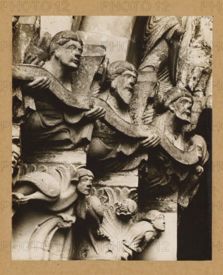 Detail of a capital of the Gnadenpforte in Bamberg Cathedral, Marienpforte, left. Capital zone Right front, album page, photograph, This photo is part of an album., number: '28.', Bamberg, 1900 - c. 1920, photographer, Bavaria, paper, baryta paper, gelatin silver print, developed gelatin silver print, height 205 mm x width 168 mm, church (exterior), Bamberg Cathedral, A close-up photograph of a carved stone relief mounted on a tan mat, showing multiple human figures in varying poses. The upper portion features three older-looking figures with carved hair and beards, two facing sideways and one turned toward the right, each holding or supporting a long, curved stone element that weaves across the composition. Below them, a crouching figure with a rounded face and stylized hair peers outward, positioned among carved architectural elements and ornamental foliage. The stone surfaces show weathering and textured patina, with deep shadows highlighting folds in drapery, carved facial features...