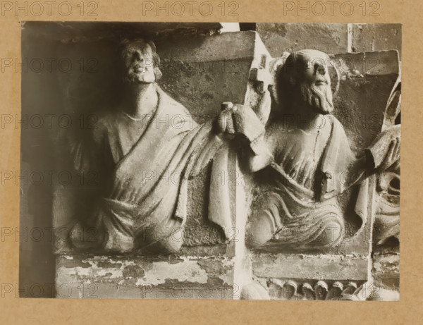 Detail of a capital of the Gnadenpforte in Bamberg Cathedral, Marienpforte, r. Kapitellzone, li vS, album page, photograph, This photo is part of an album., number: '29.', Bamberg, 1900 - c. 1920, photographer, Bavaria, paper, baryta paper, gelatin silver print, developed gelatin silver print, height 162 mm x width 221 mm, church (exterior), Bamberg Cathedral, A cropped black-and-white photograph of two carved stone figures seated side by side, each wearing draped garments with visible folds; the figure on the left has a rounded face and short hair, the figure on the right has a beard and a halo-like circular carving behind the head; both figures rest on a textured stone background with a vertical divider between them and a horizontal ledge below that shows chipped and weathered surfaces.