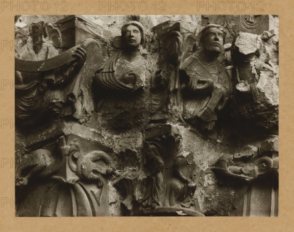 Detail of a capital of the Gnadenpforte in Bamberg Cathedral, Marienpforte, r. Kapitellzone, rv SO., album page, photograph, This photo is part of an album., number: '33.', Bamberg, 1900 - c. 1920, photographer, Bavaria, paper, baryta paper, gelatin silver print, developed gelatin silver print, height 168 mm x width 220 mm, church (exterior), Bamberg Cathedral, A close-up view of a weathered stone relief shows several carved human figures and decorative elements arranged on a rough background. Two larger upper figures face forward with detailed facial features, draped garments, and stylized wings or flowing cloth around their shoulders, one with a hand raised near the chest and the other with a hand extended to the side. Below and around them are smaller, partially damaged figures and ornamental shapes, including curved drapery, fragmented architectural details, and rough, chipped stone surfaces. The entire scene is monochromatic with strong contrasts between smooth carved areas and co...