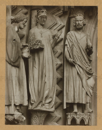 Adamspforte, left wall of the North., Detail of the Adamspforte in Bamberg Cathedral, album page, photograph, This photo is part of an album., number: '36.', Bamberg, 1900 - c. 1920, photographer, Bavaria, paper, baryta paper, gelatin silver print, developed gelatin silver print, height 210 mm x width 163 mm, church (exterior), Bamberg Cathedral, Three stone figures carved in relief stand side by side against a textured architectural background, each wearing long draped garments and ornate crowns; the left figure leans slightly forward with curly hair and extends hands holding a small sculpted object, the central figure faces forward with one hand raised and wears a necklace while holding a model-like structure, and the right figure has a beard and holds a spherical object in one hand with the other hand resting on drapery, all figures show detailed folds in their robes and stand on carved pedestals with geometric and floral motifs.