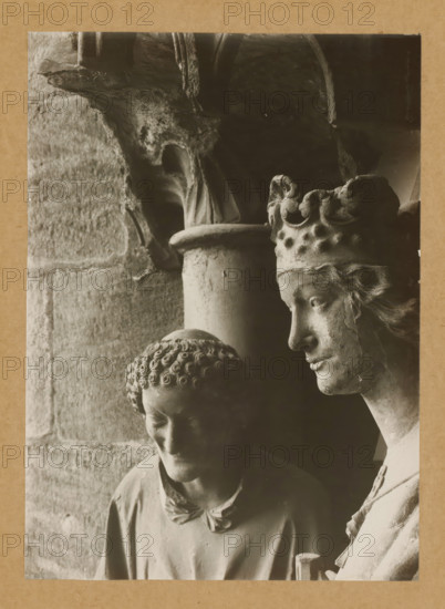 Adamspforte, Stephen and Kunigunde (Heads), Detail of the Adamspforte in Bamberg Cathedral, album page, photograph, This photo is part of an album., number: '38.', Bamberg, 1900 - c. 1920, photographer, Bavaria, paper, baryta paper, gelatin silver print, height 223 mm x width 161 mm, Person, Stephen (martyr), church (exterior), Bamberg Cathedral, A close-up monochrome photograph of two stone sculptures positioned side by side against a textured stone wall and a dark background, showing the right-profile of a crowned figure with carved hair, a serene facial expression, and a detailed ornate crown, next to a lower-headed figure with tightly curled hair, downcast eyes, and a simple draped garment, with visible weathering and surface texture on both sculptures and architectural stone elements above and behind them.