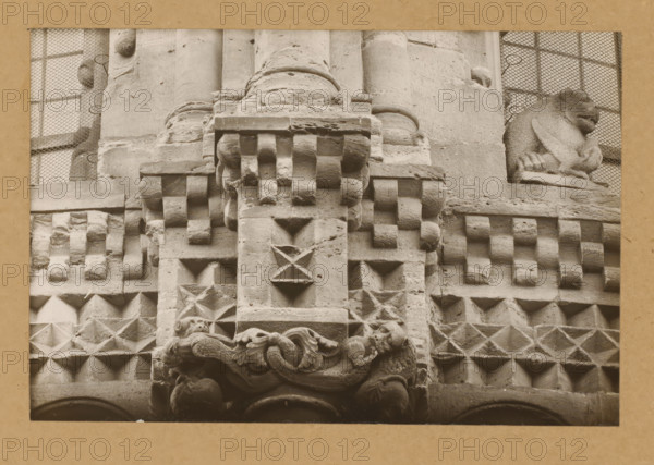 East Church, 1. Frieze 3., View of the sculpture of a frieze of Bamberg Cathedral, album page, photograph, This photo is part of an album., number: '5.', Bamberg, 1900 - c. 1920, photographer, Bavaria, paper, baryta paper, gelatin silver print, height 141 mm x width 219 mm, church (exterior), Bamberg Cathedral, A close-up of a carved stone façade showing geometric protruding blocks and pyramidal shapes, a central vertical stone column with sculpted decorative elements including rounded blocks and recessed diamond and triangular recesses, ornate carved scrollwork and foliage near the bottom, a sculpted animal figure perched on a corner ledge on the right, and mesh-covered window openings visible in the upper corners.