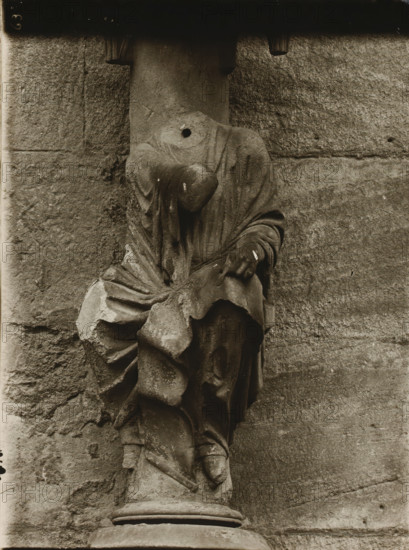Detail of the Fürstenportal in the cathedral of Bamberg, Adamspforte, Johannes vN, album page, photograph, This photo is part of an album., number: '63', inscription: 'Kunsthist. Seminar, Marburg (Lahn), Photogr. Abteilung, Vervielfältigung, untersagt.', Bamberg, 1900 - c. 1920, photographer, Bavaria, paper, baryta paper, gelatin silver print, developing gelatin silver print, height 238 mm x width 180 mm, church (exterior), Bamberg Cathedral, A weathered stone sculpture of a draped figure mounted against a rough stone wall, showing folded and flowing fabric over a torso and limbs, with a rounded protrusion where a head might be and a circular hole in the upper chest area; the sculpture rests on a cylindrical stone plinth and displays chips, discoloration, and surface wear across the folds and edges, while the background consists of textured masonry blocks with visible joints and worn surfaces.