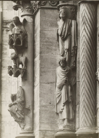 Adamspforte, Evangelisten u. Statuensäule li.lvN, Detail of the Fürstenportal in Bamberg Cathedral, album page, photograph, This photo is part of an album., inscription: 'Kunsthist. Seminar, Marburg (Lahn), Photogr. Abteilung, Vervielfältigung, untersagt.', number: '64', Bamberg, 1900 - c. 1920, photographer, Bavaria, paper, baryta paper, gelatin silver print, developing gelatin silver print, height 237 mm x width 176 mm, church (exterior), Bamberg Cathedral, A close view of carved stone figures and architectural details on a building façade, showing sculpted human figures in draped clothing with carved facial features and beards, a seated figure holding an object, a small animal figure standing on a decorative ledge with miniature arch motifs, ornate column capitals with floral and scroll patterns, a fluted column with diagonal chevron carving, and blocks of masonry with visible joints and weathering.
