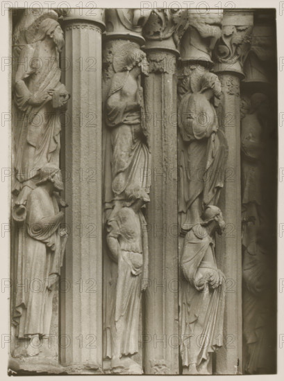 Detail of the Fürstenportal in Bamberg Cathedral, Evangelistensäule Stat. li. 2.3.4.vN, album page, photograph, This photo is part of an album., inscription: 'Kunsthist. Seminar, Marburg (Lahn), Photogr. Abteilung, Vervielfältigung, untersagt.', inscription: '67, Bamberg', Bamberg, 1900 - c. 1920, photographer, Bavaria, paper, baryta paper, gelatin silver print, developing gelatin silver print, height 236 mm x width 179 mm, church (exterior), Bamberg Cathedral, A close view of a stone architectural relief showing a row of tall fluted columns with carved human figures standing in recessed niches between them, each figure draped in flowing robes with visible folds and varying head coverings or hairstyles; some faces are in profile and others angled, with hands positioned near chests or holding drapery, and smaller figures or heads appear at shoulder or knee level of larger figures; the capitals above the columns feature carved foliage and decorative motifs, and the stone surface shows su...