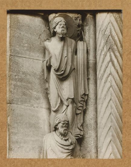 Detail of the Fürstenportal in Bamberg Cathedral, Evangelistensäule Apostel li.lvNW., album page, photograph, This photo is part of an album., number: '71', Bamberg, 1900 - c. 1920, photographer, Bavaria, paper, baryta paper, gelatin silver print, developed gelatin silver print, height 208 mm x width 162 mm, church (exterior), Bamberg Cathedral, A grayscale photograph shows two stone sculpted figures mounted on a textured wall, one positioned above the other; the upper figure wears draped robes and a halo, has a beard and long hair, and holds a fold of cloth at chest level, while the lower figure also has a beard and draped garments and appears with a turned head; to the right of the figures is a vertical carved column with a chevron pattern, and the surrounding stone surface shows weathered texture and horizontal joint lines, all framed by a mottled rectangular border.