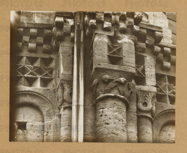 East Church, 1. Frieze 5., View of the sculpture of a frieze of Bamberg Cathedral, album page, photograph, This photo is part of an album., number: '7.', Bamberg, 1900 - c. 1920, photographer, Bavaria, paper, baryta paper, gelatin silver print, developed gelatin silver print, height 167 mm x width 211 mm, church (exterior), Bamberg Cathedral, A close view of aged stone architectural details showing carved blocks and columns with weathered surfaces, patterned geometric carvings including raised square and diamond shapes, rounded column shafts with fluted and pockmarked textures, a smooth vertical metal pipe running down the center, and fragments of ornamental molding and carved rosettes, all in a monochrome sepia tone with a plain border around the image.