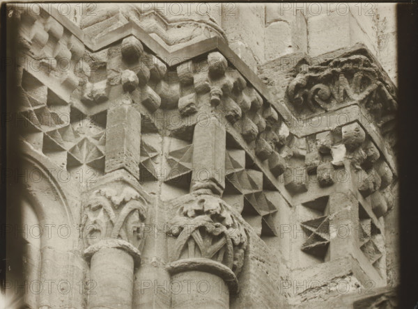 East Church, 1. Frieze 6., View of the sculpture of a frieze of Bamberg Cathedral, album page, photograph, This photo is part of an album., inscription: 'Kunsthist. Seminar, Marburg (Lahn), Photogr. Abteilung, Vervielfältigung, untersagt.', Bamberg, 1900 - c. 1920, photographer, Bavaria, paper, baryta paper, gelatin silver print, developed gelatin silver print, height 178 mm x width 235 mm, church (exterior), Bamberg Cathedral, A close-up view of a carved stone surface showing architectural details including rounded columns with smooth shafts and molded capitals, geometric star-like recessed patterns carved into square panels, rows of small rounded boss-like projections, and intricately carved foliage motifs on a corner capital; the stone has a weathered texture with visible tool marks and chips, and the image is monochrome with a soft vignette around the edges.