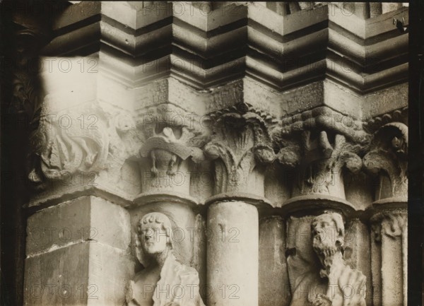 Detail of the Fürstenportal in Bamberg Cathedral, Fürstentor, Kapitelle r. über 1.2.v.NO., album page, photograph, This photo is part of an album., inscription: 'Bamberg, 83', inscription: 'Kunsthist. Seminar, Marburg (Lahn), Photogr. Abteilung, Vervielfältigung, untersagt.', Bamberg, 1900 - c. 1920, photographer, Bavaria, paper, baryta paper, gelatin silver print, developing gelatin silver print, height 177 mm x width 238 mm, church (exterior), Bamberg Cathedral, A close view of a stone architectural detail showing carved capitals and columns with layered mouldings above, featuring sculpted foliage motifs and animal-like forms, with two carved human figures set into vertical niches beside the columns, one with a youthful face and short curly hair and the other with a bearded face and draped clothing, all rendered in weathered pale stone with areas of shadow and darker grime and a bright washed area near the upper left.